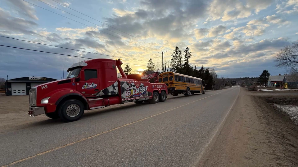Au coucher du soleil, la dépanneuse lourde de Remorquage Gilbert remorque un autobus scolaire jaune sur une route de village en Mauricie