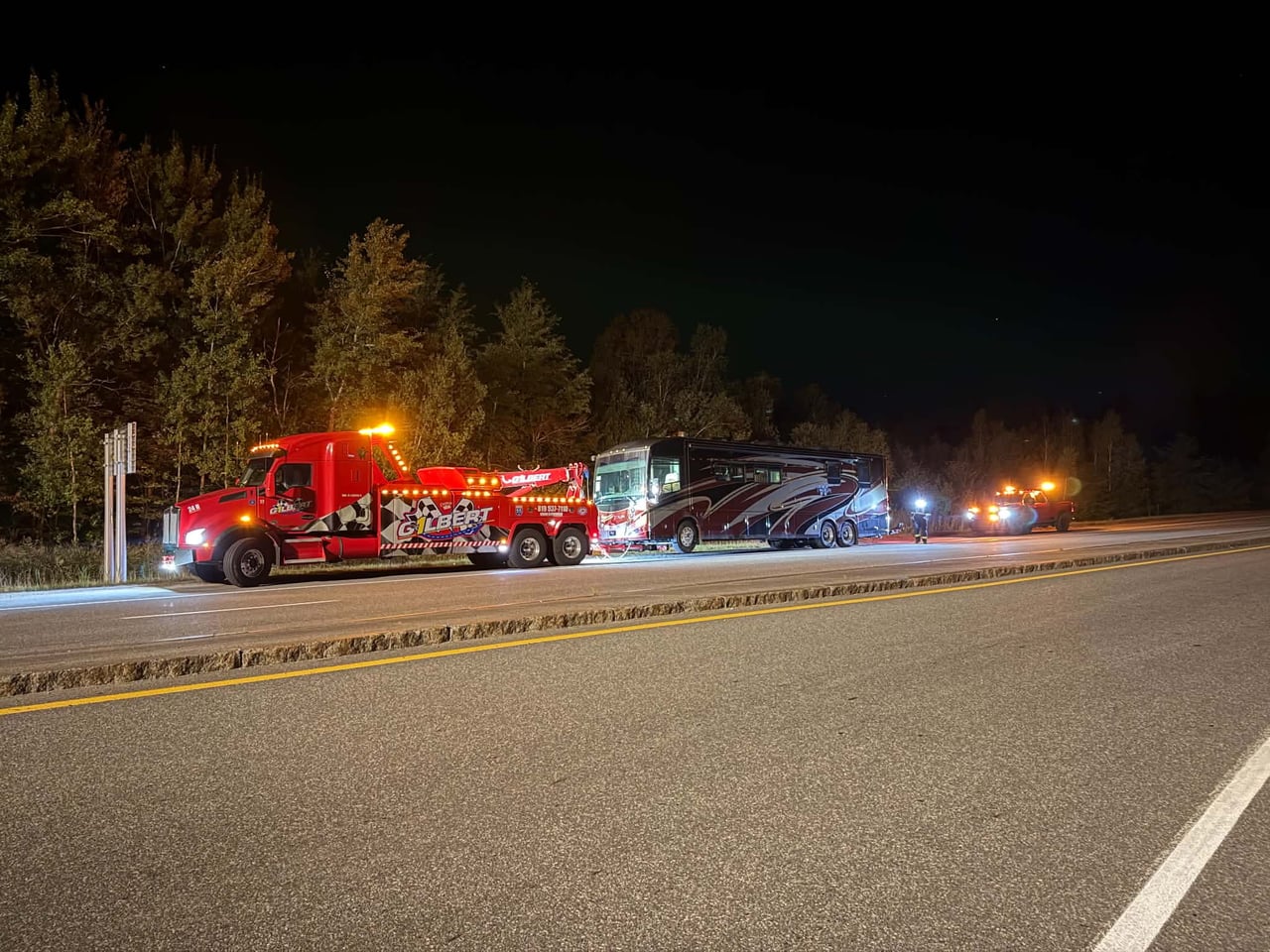 De nuit, une dépanneuse lourde rouge de Remorquage Gilbert, gyrophares allumés, remorque un autocaravane sur l’accotement d’une route de la Mauricie