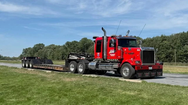 Transport de machinerie : tracteur routier Western Star rouge de Remorquage Gilbert attelé à un fardier surbaissé à trois essieux, en bordure de route